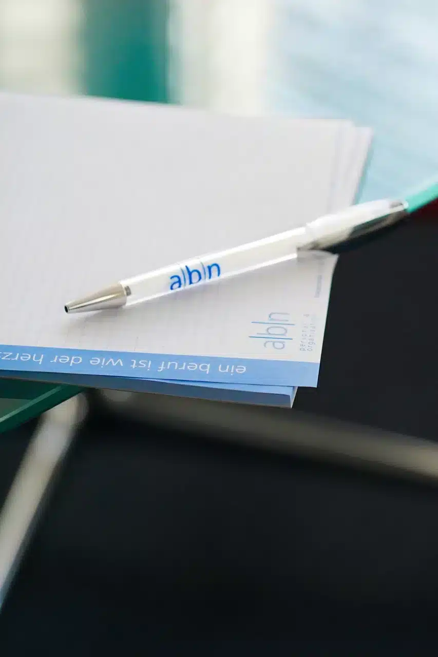 organisation-optimierung-abn Kugelschreiber und Notizblock mit abn-Logo auf einem Glastisch