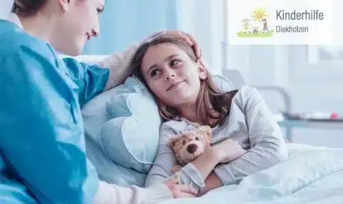 Mädchen im Krankenhausbett erhält Trost durch eine Betreuerin mit Teddybär
